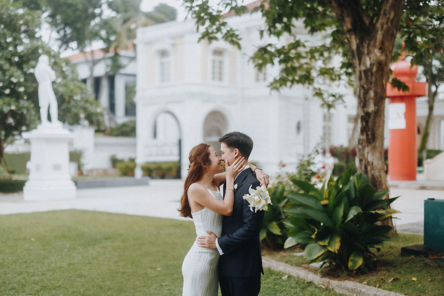 Couple poses romantically on a lawn in front of a white colonial-style building.