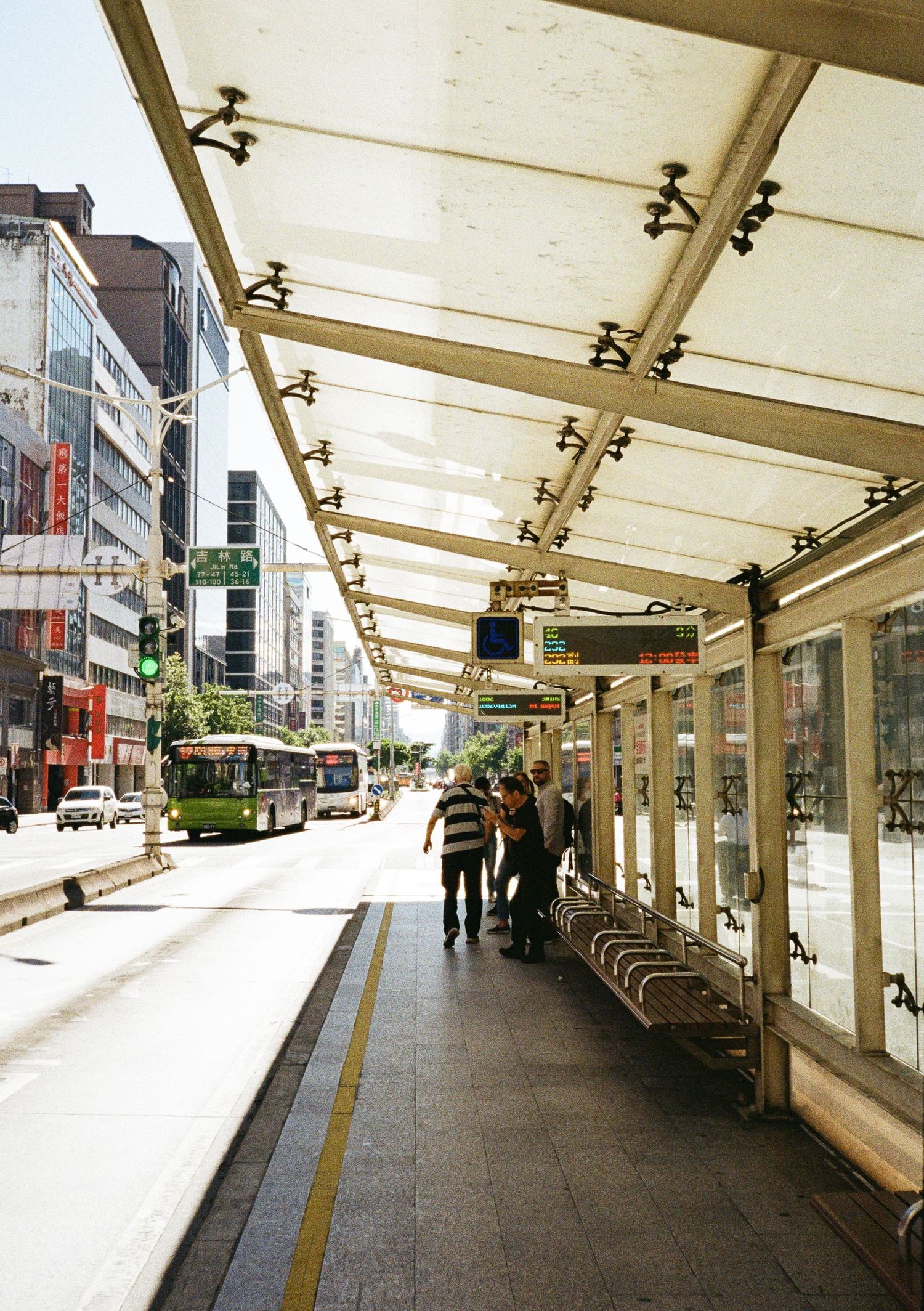 Modern covered bus stop with illuminated ceiling panels along a city street with buildings and transit.