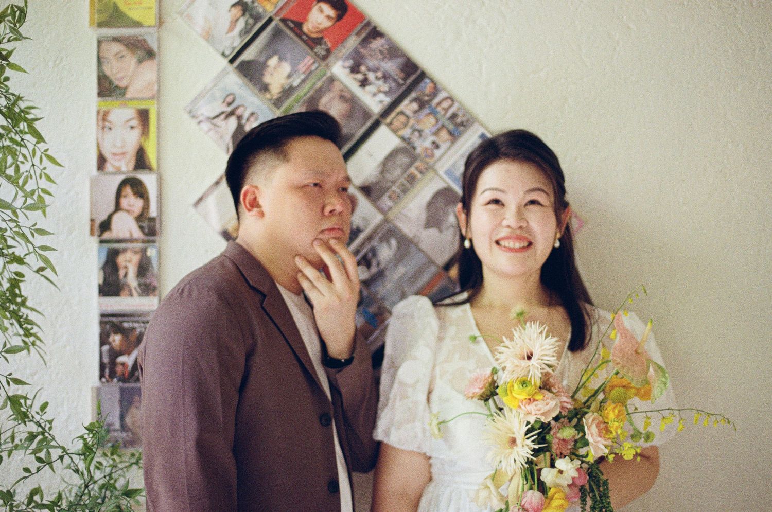 A joyful couple posing together in front of a wall decorated with photographs holding yellow flowers.