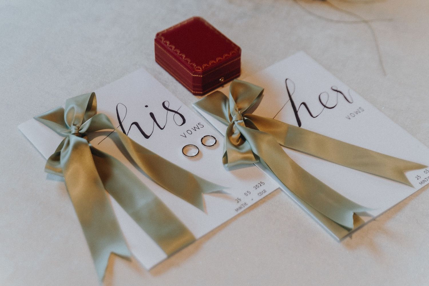 Wedding rings and stationery with sage green ribbons and a red velvet ring box.