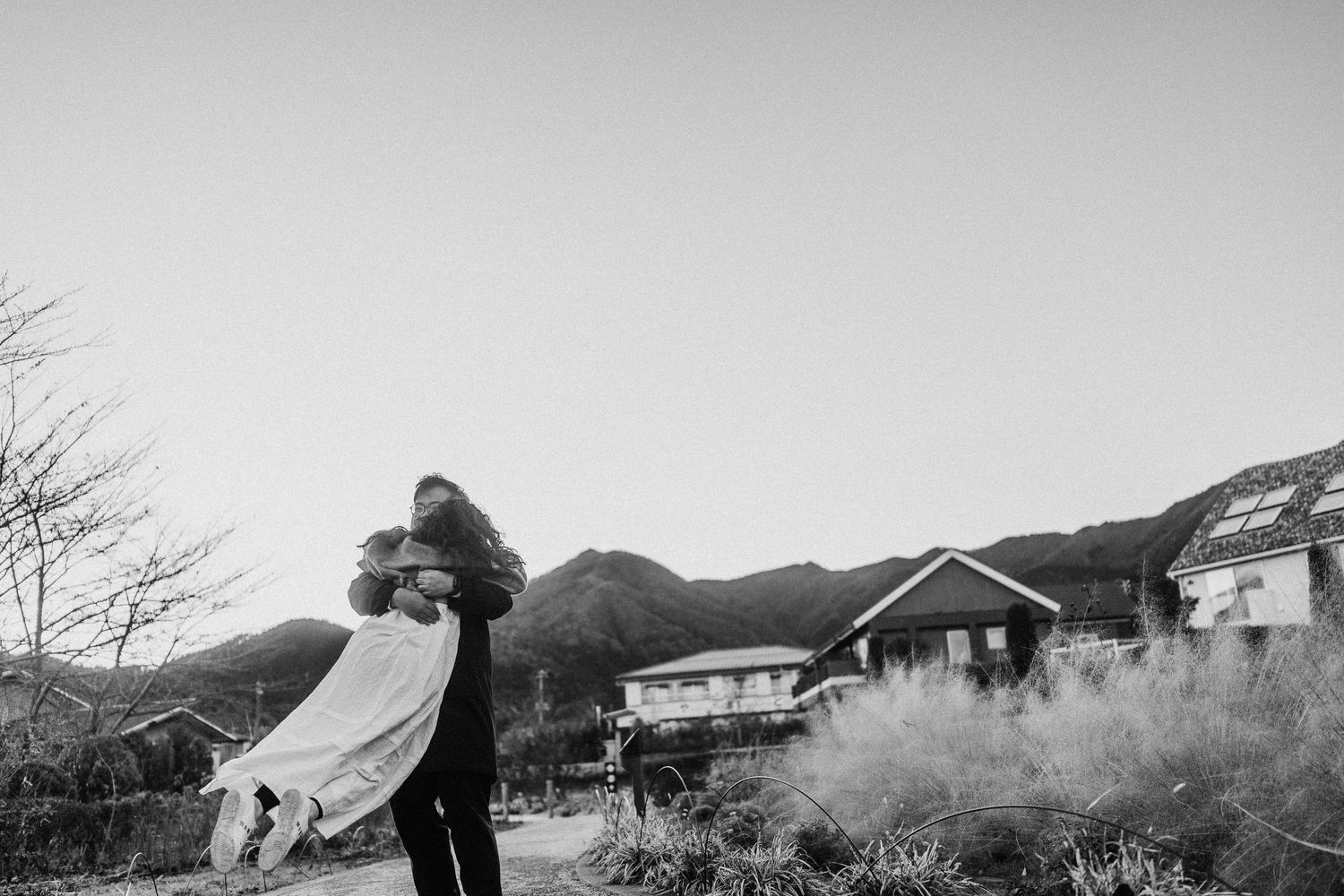 A black and white portrait of two people embracing with mountain scenery in the background.