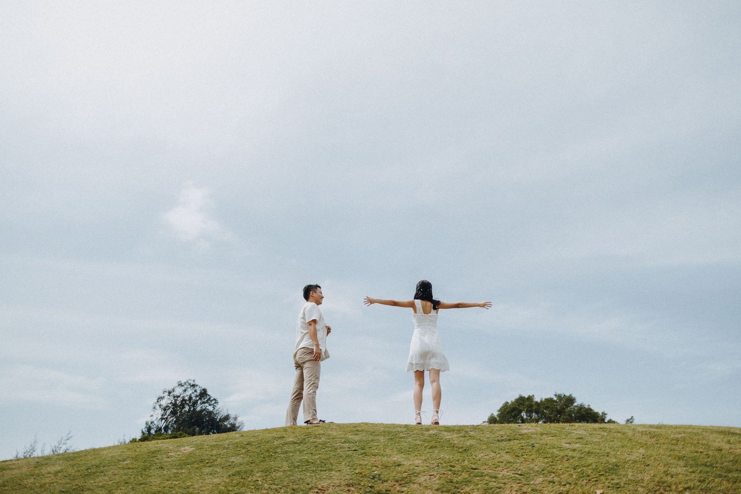 Two people stand on a hilltop with arms outstretched against a cloudy sky.