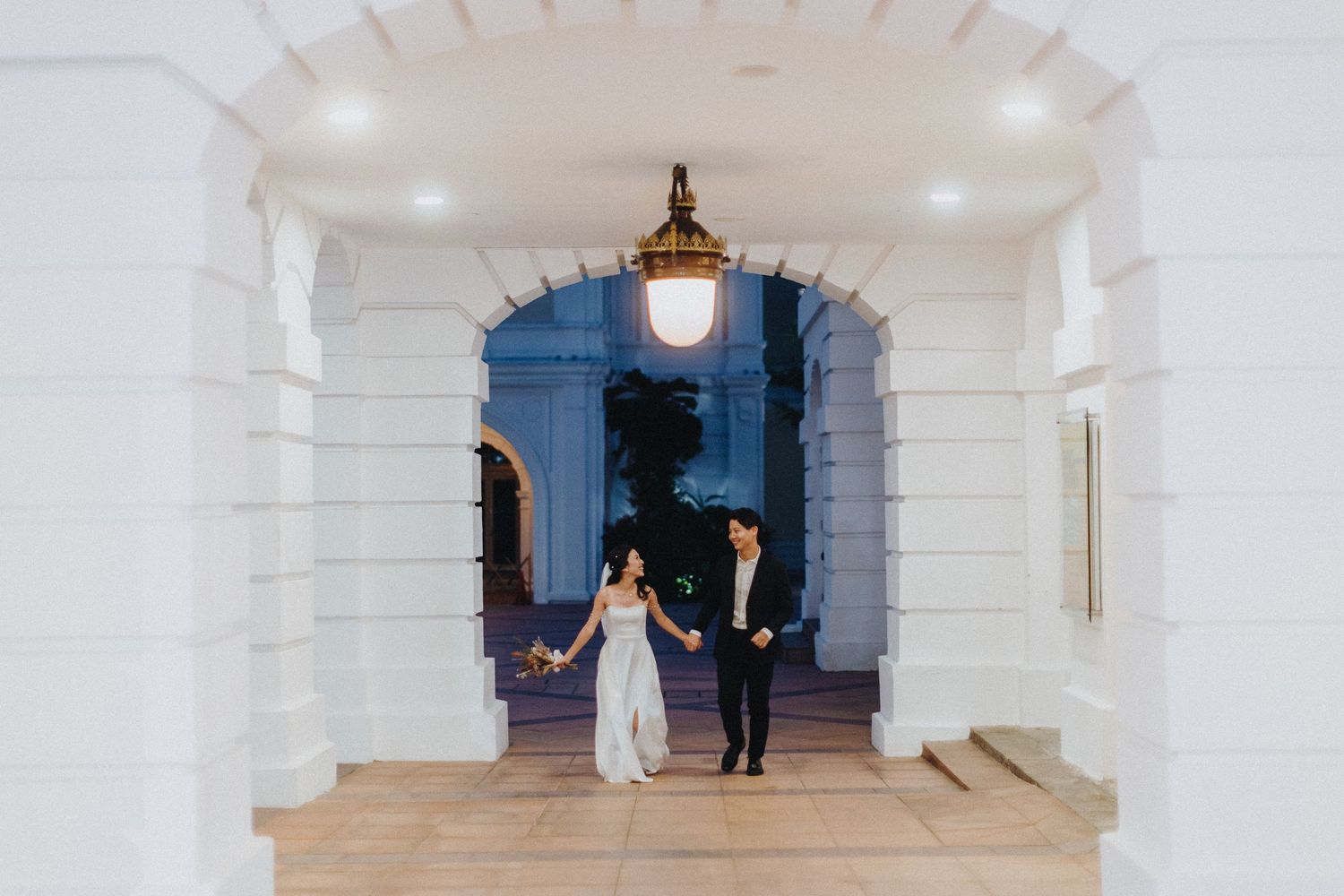 Two people walking through an elegant white arched corridor with romantic lighting.