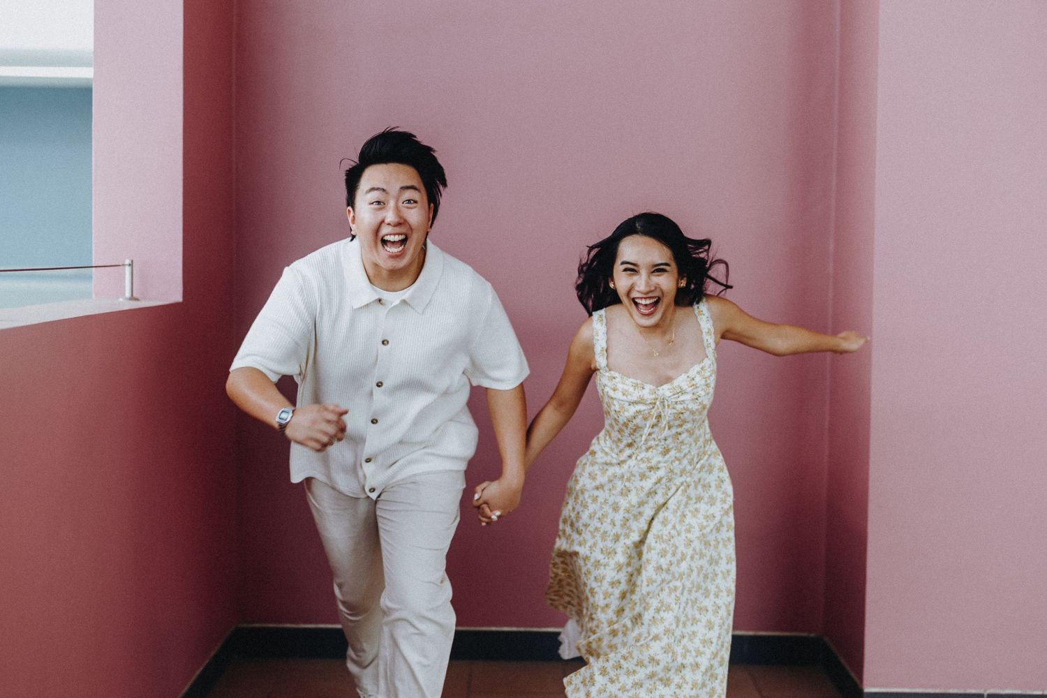 Couple displaying animated expressions against a pink wall in contrasting outfits.