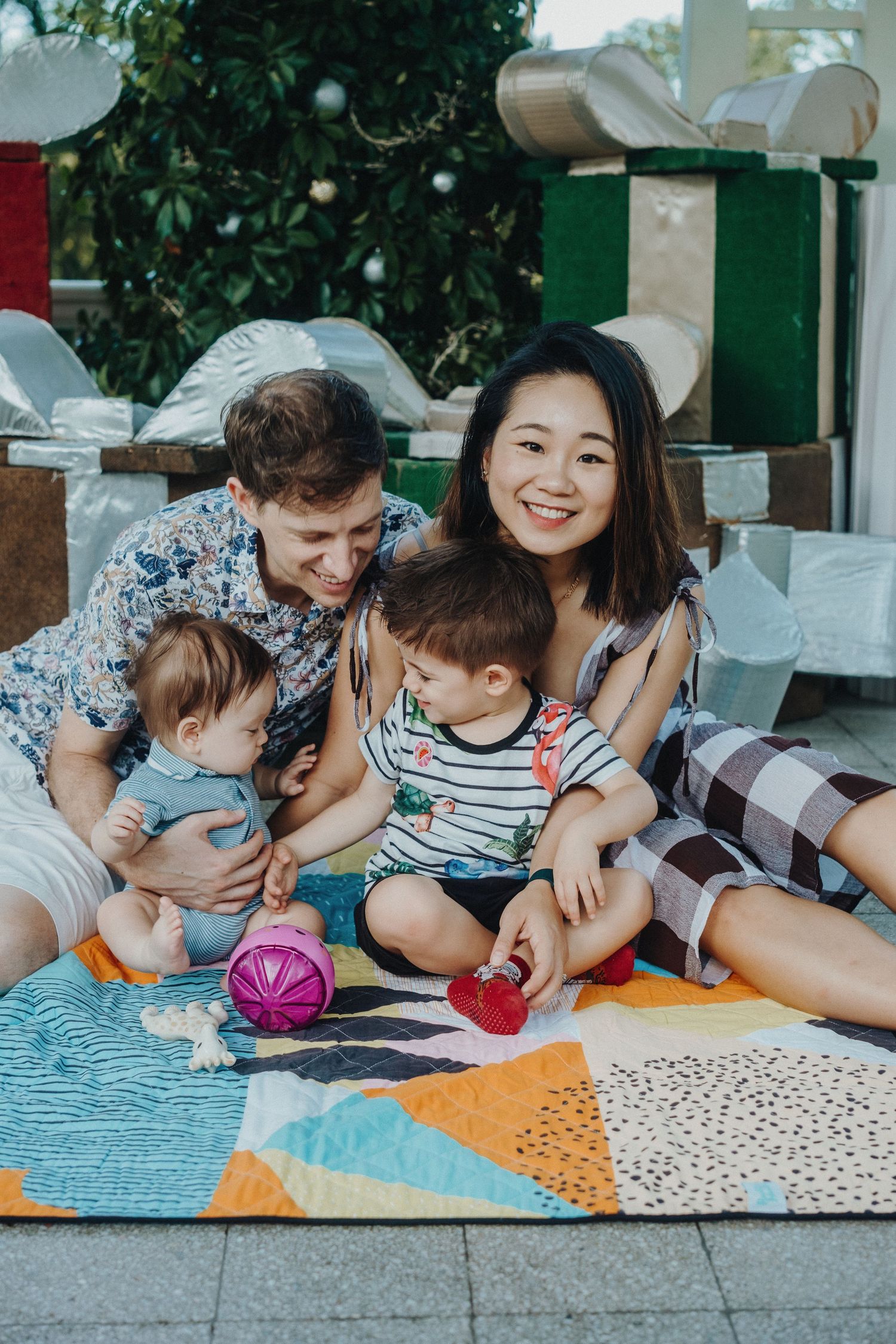 A family sits together on a colorful patchwork blanket with toys during an outdoor playdate.