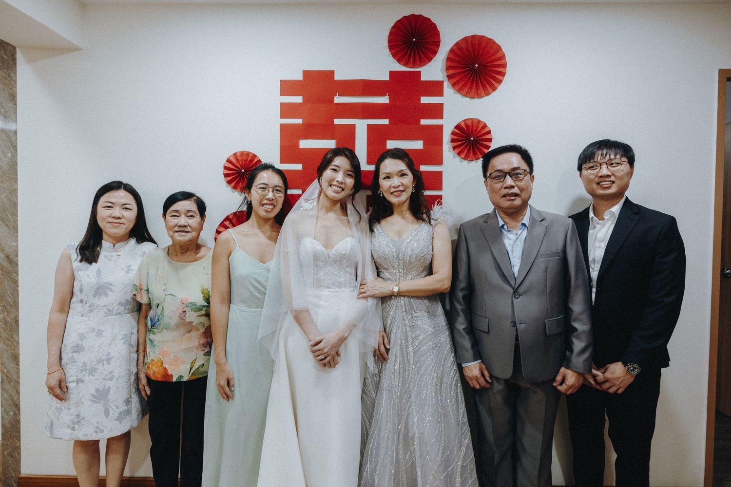 Group photo at wedding tea ceremony with decorative red wall and Chinese happiness symbol in background.
