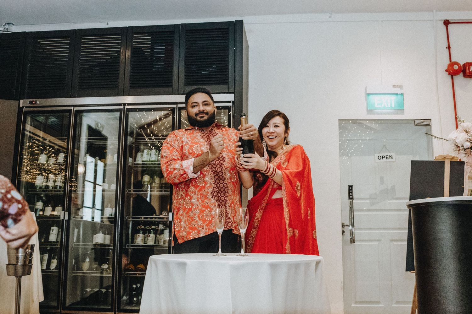 A couple in traditional Indian and batik attire stands together at a wedding reception table.