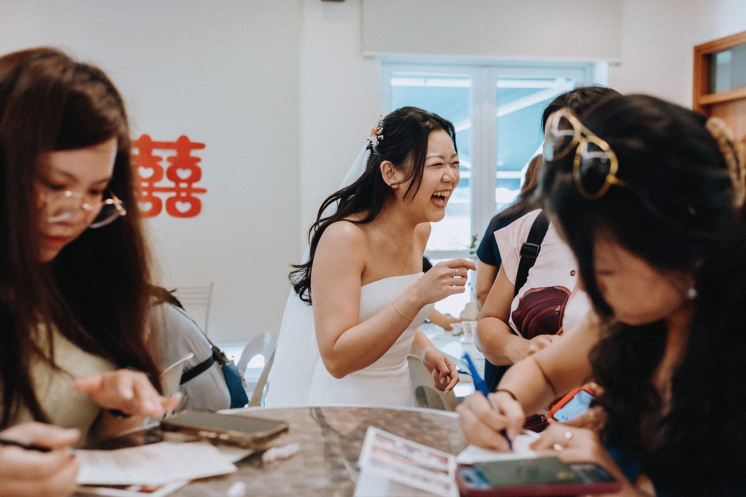 Asian bride in strapless white dress laughs with bridesmaids at table beneath red double happiness symbol.