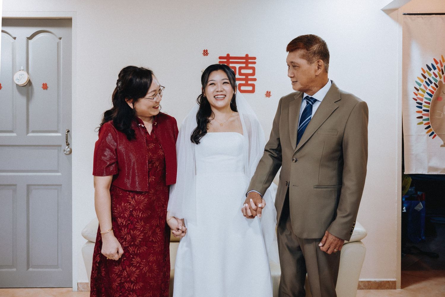 Bride in white dress stands smiling with parents in formal attire near Chinese double happiness symbol decor.