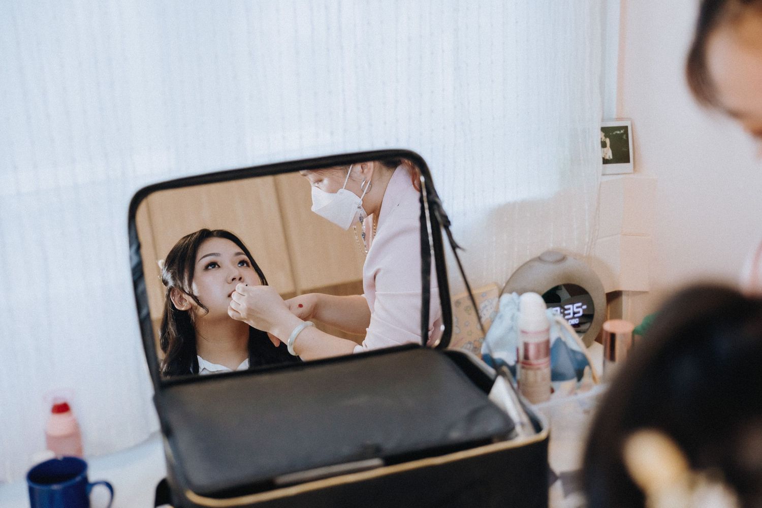 A bride gets her makeup applied by a masked artist, reflected in a professional vanity mirror.