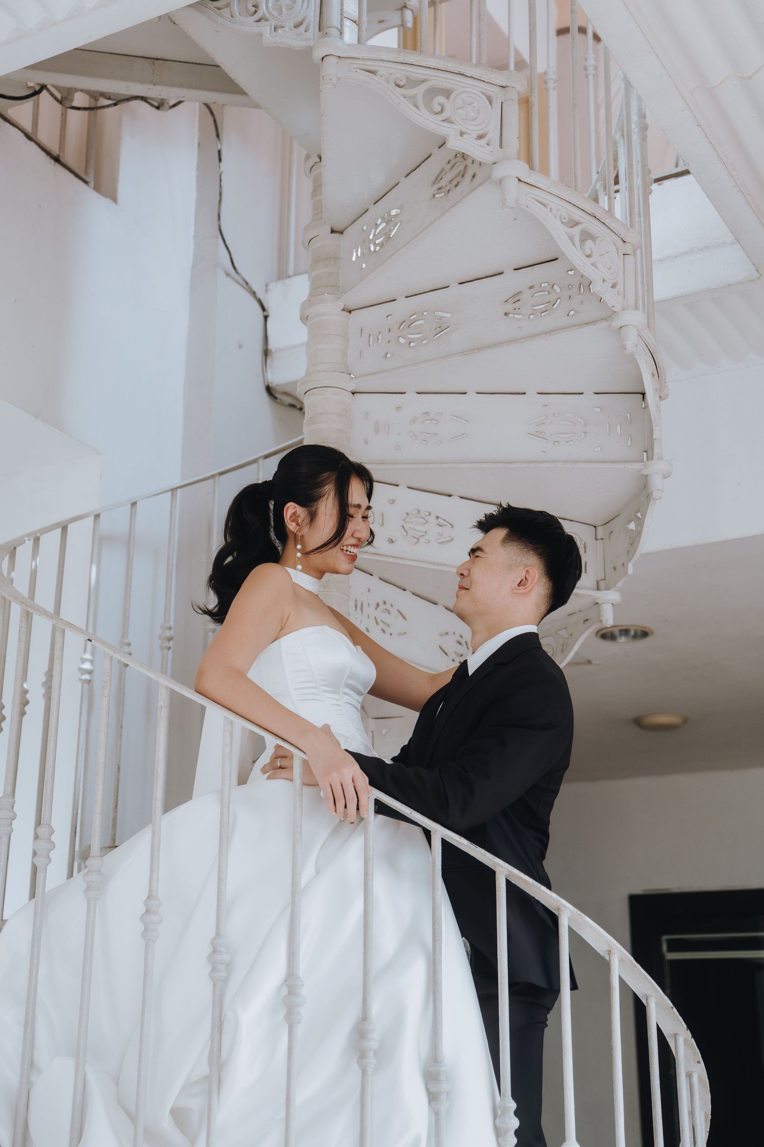 A bride and groom share a romantic moment on a white spiral staircase, dressed in wedding attire.