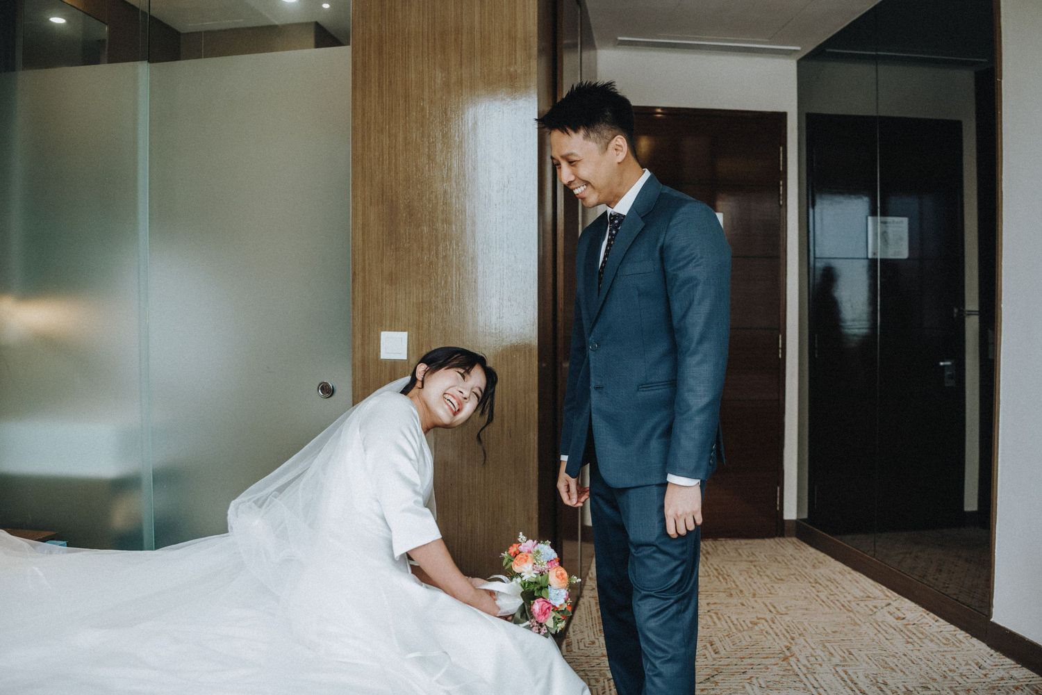 Bride in white gown sits on bed holding bouquet while groom in navy suit stands nearby in elegant hotel room.