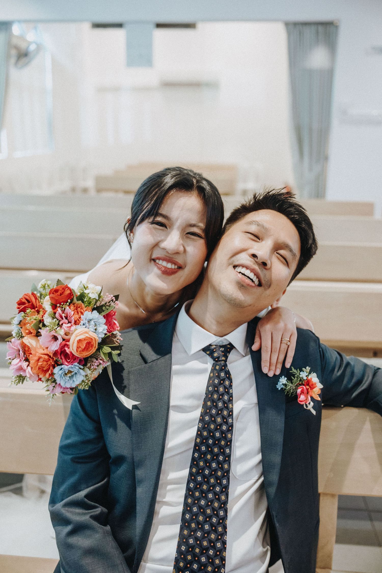 Happy Asian bride and groom smiling together in church pew, bride holding colorful bouquet of flowers.
