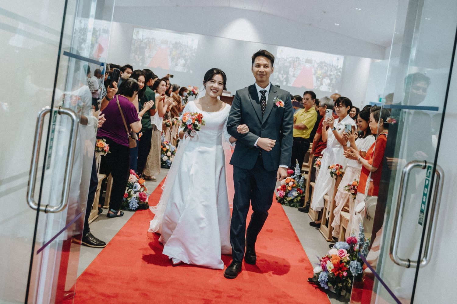 A bride and groom walk down a red carpet aisle surrounded by guests at their wedding ceremony.