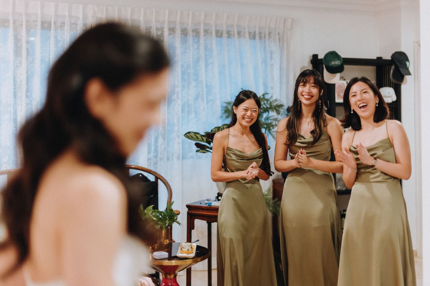 Three bridesmaids in olive green satin dresses laughing at bride during wedding morning preparations.