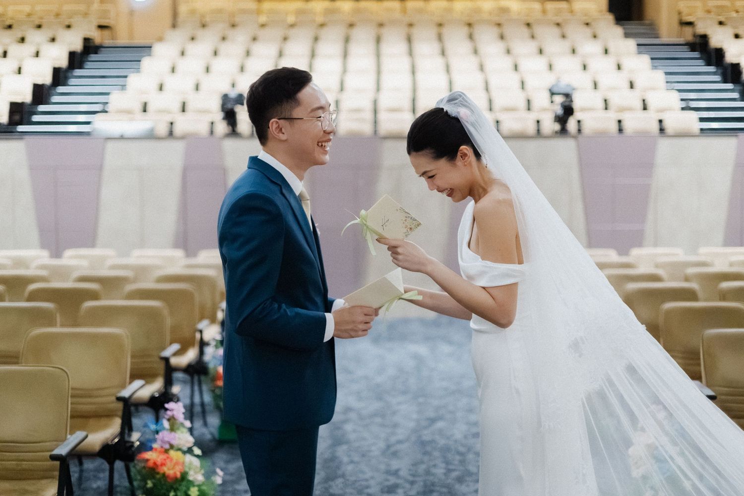 Asian bride reads vows from a card while groom watches lovingly during wedding ceremony in an auditorium venue.