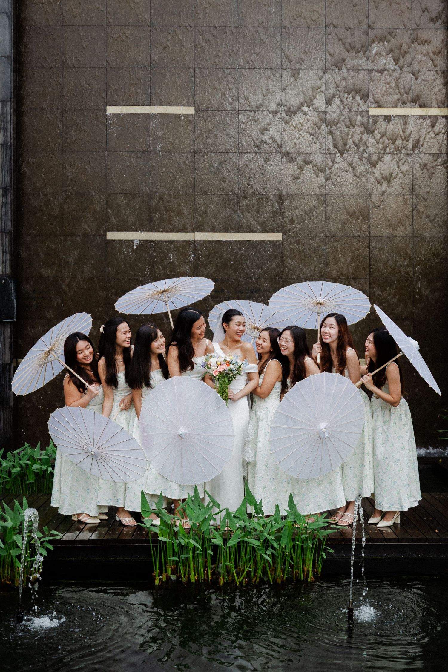 Bride surrounded by bridesmaids in mint dresses holding white paper parasols near decorative fountain at outdoor wedding venue.