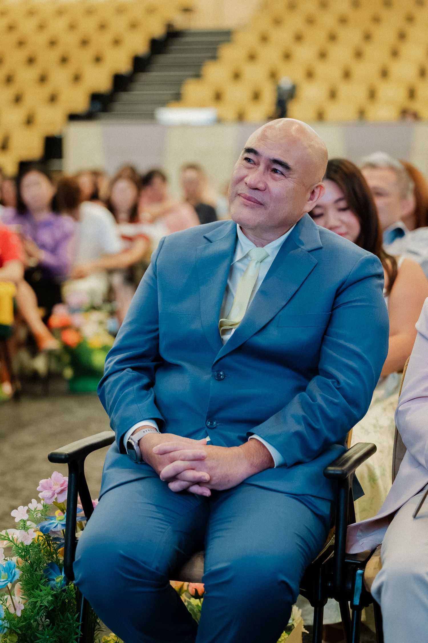 Bald man in blue suit smiling while seated at a wedding ceremony with guests in background.