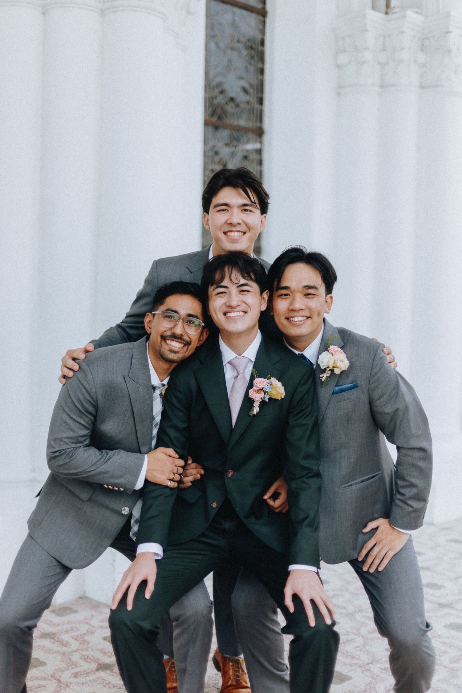 Smiling groom in dark suit surrounded by three groomsmen in grey suits posing together outside white building.