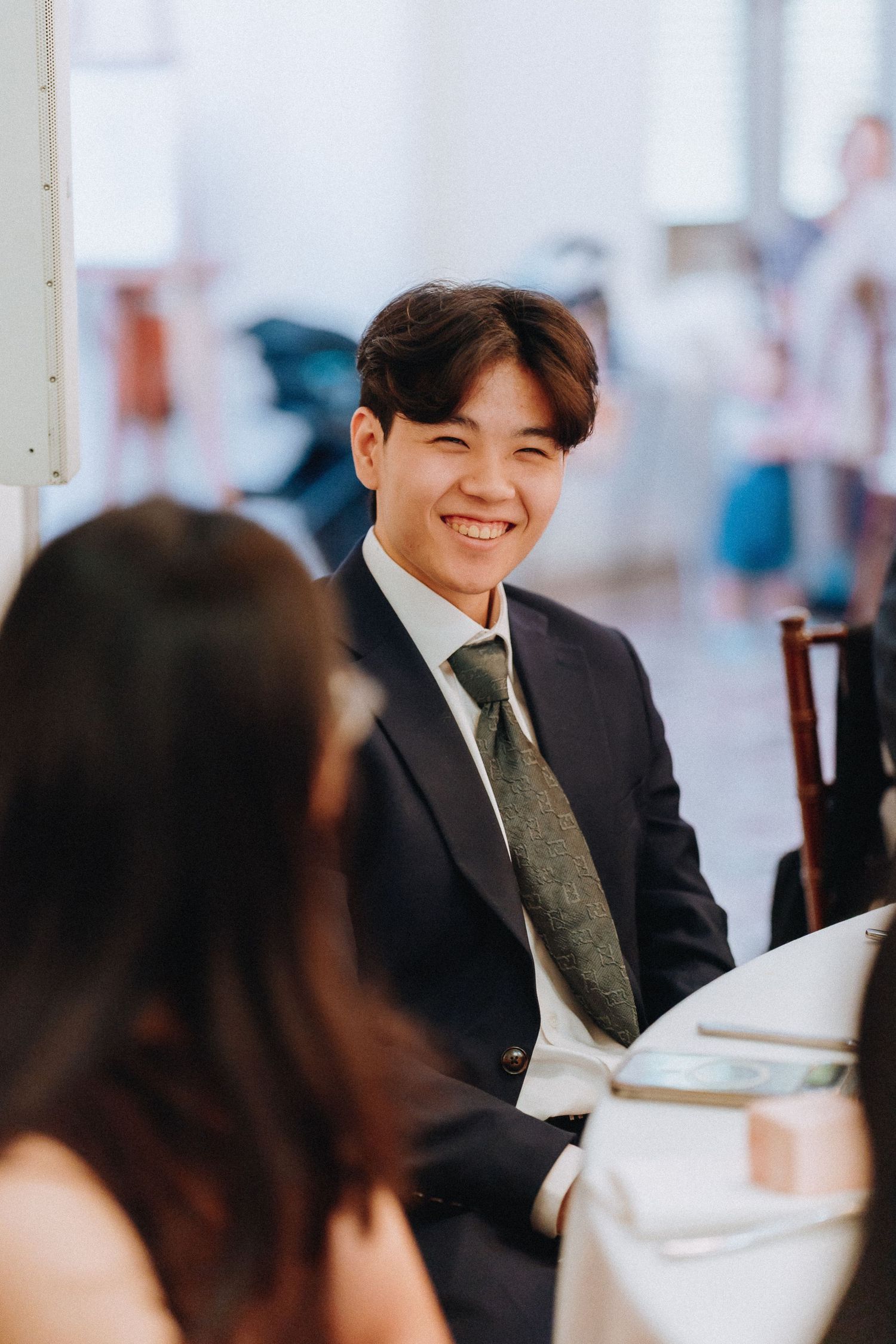 Smiling young man in a dark suit and patterned tie seated at a wedding reception table.