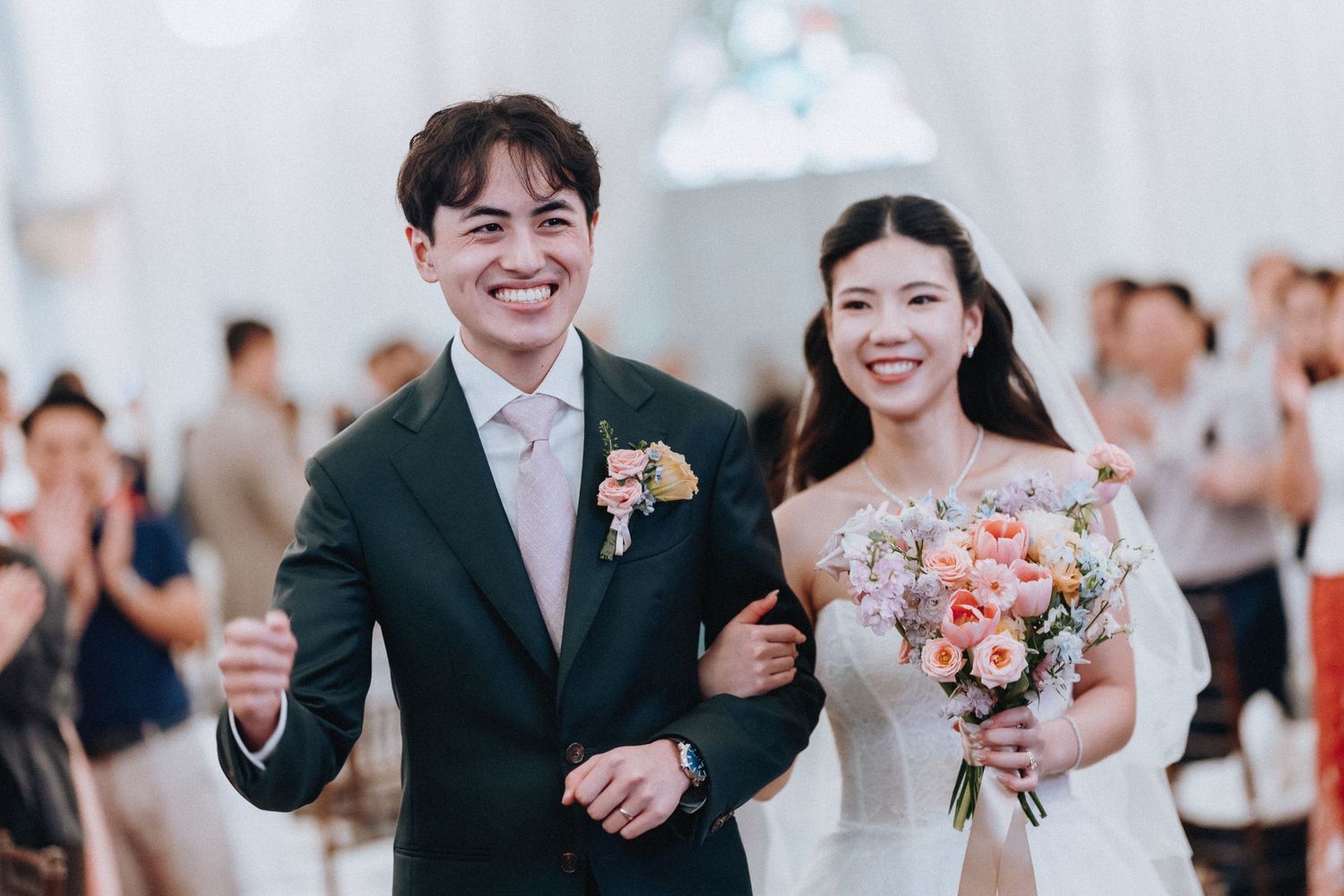 Happy couple walking down the aisle at their wedding ceremony, groom in dark suit, bride in white gown holding pink bouquet.