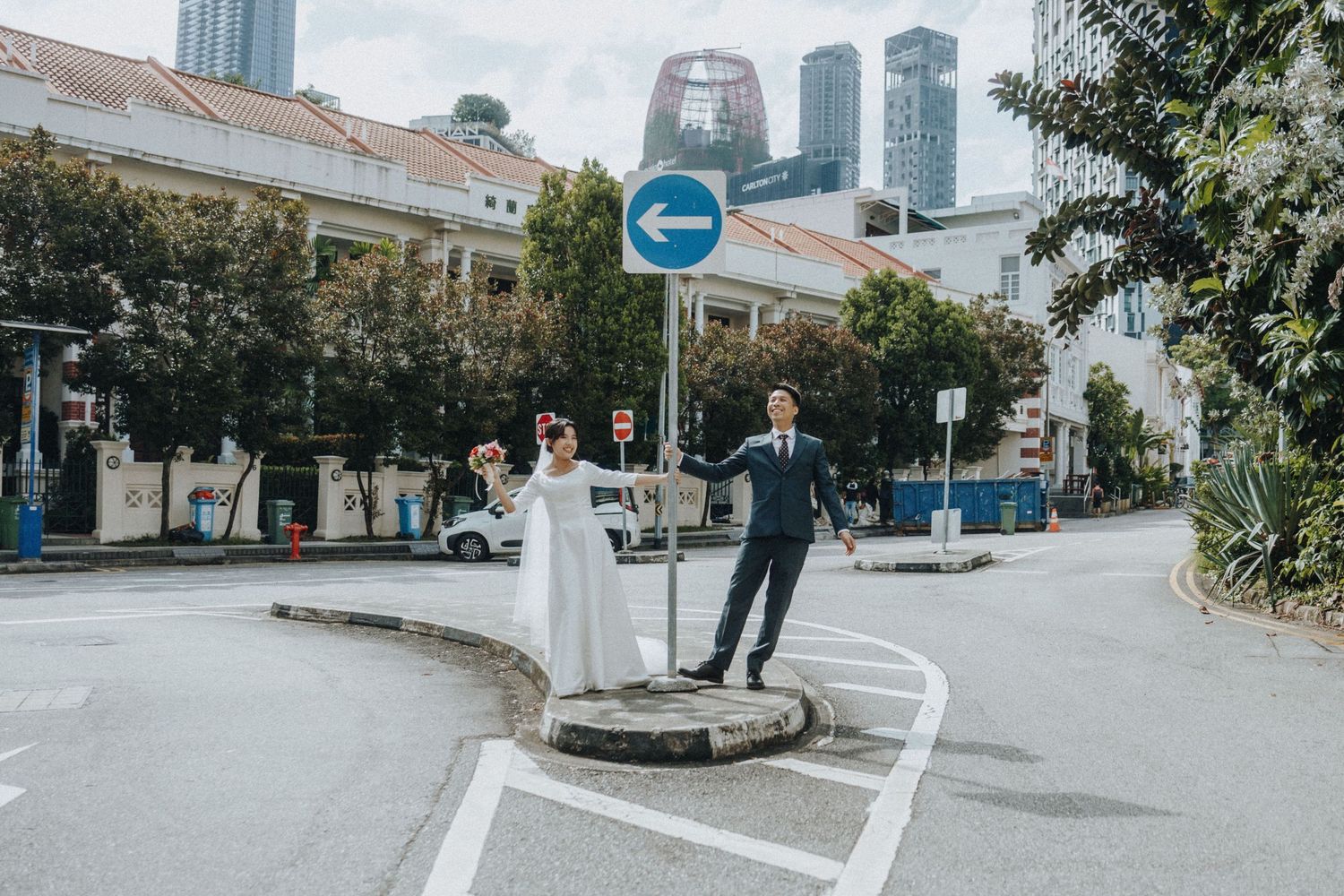 A joyful couple in wedding attire celebrates on a street corner with city buildings in the background.