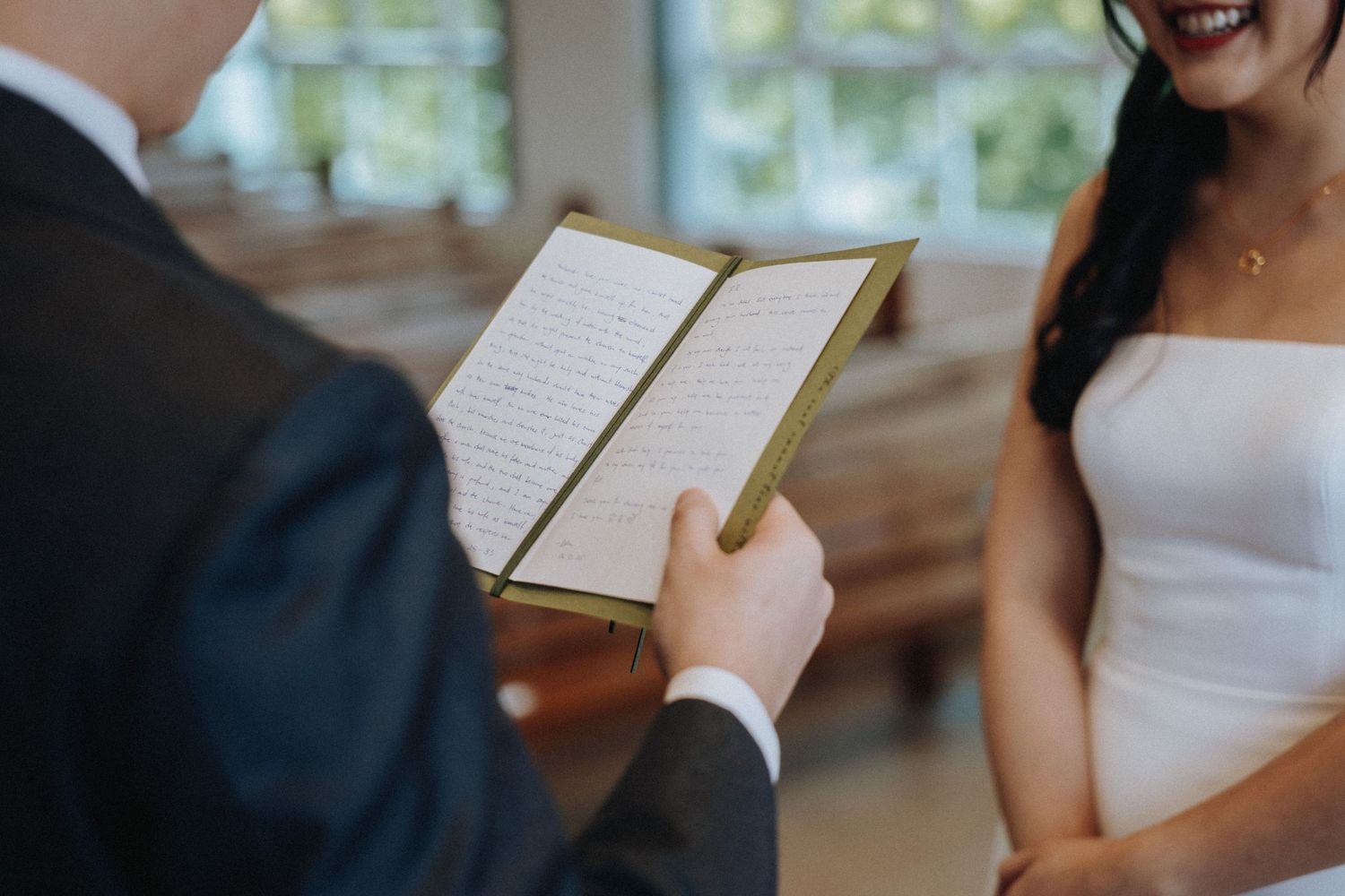 Groom reads wedding vows from a gold booklet as smiling bride listens during church ceremony.
