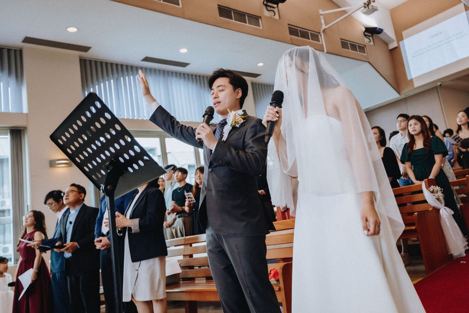 Groom sings with microphone raised hand at church wedding ceremony beside bride in white veil.