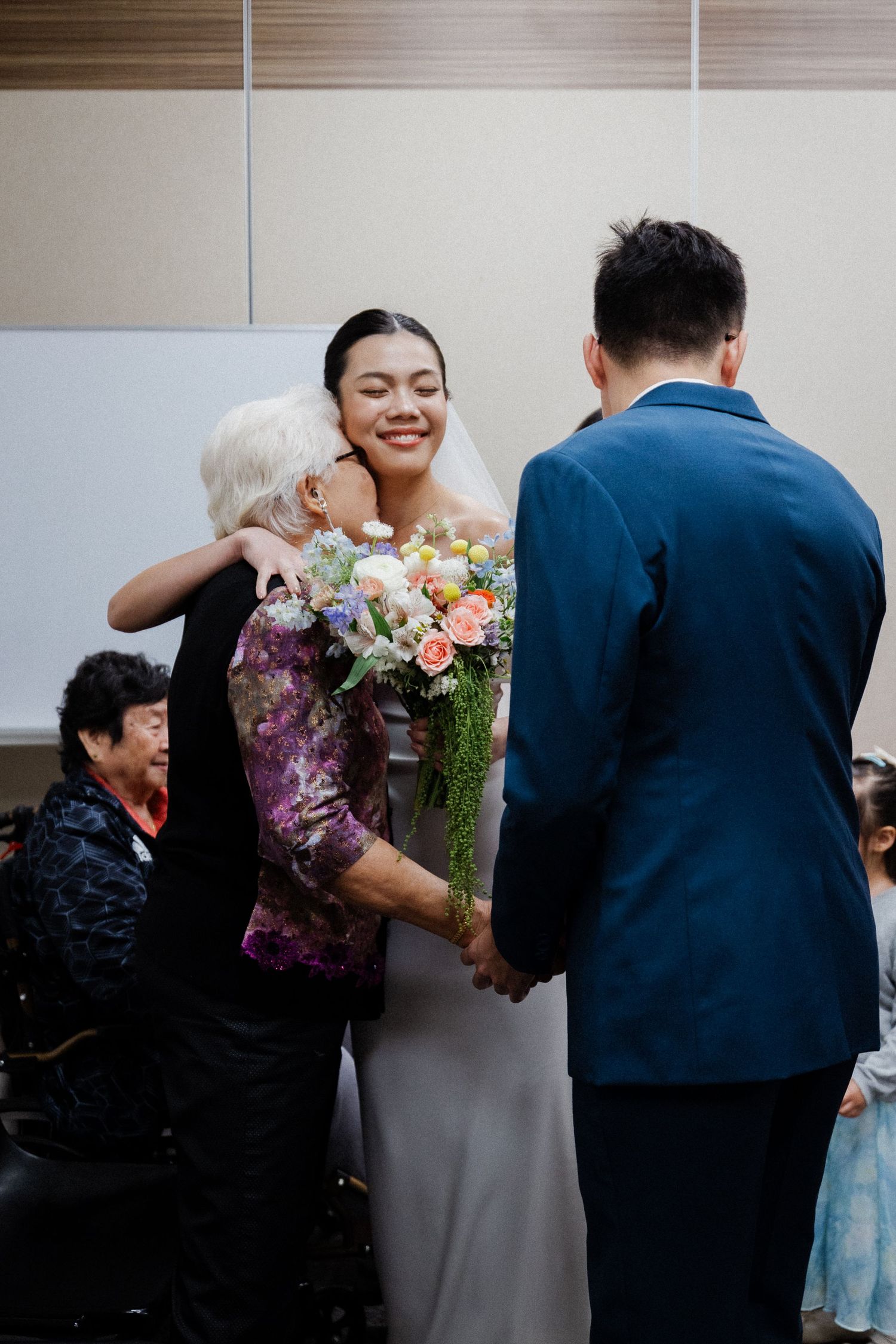 Bride in white veil embraces elderly guest while groom in blue suit holds her hand.