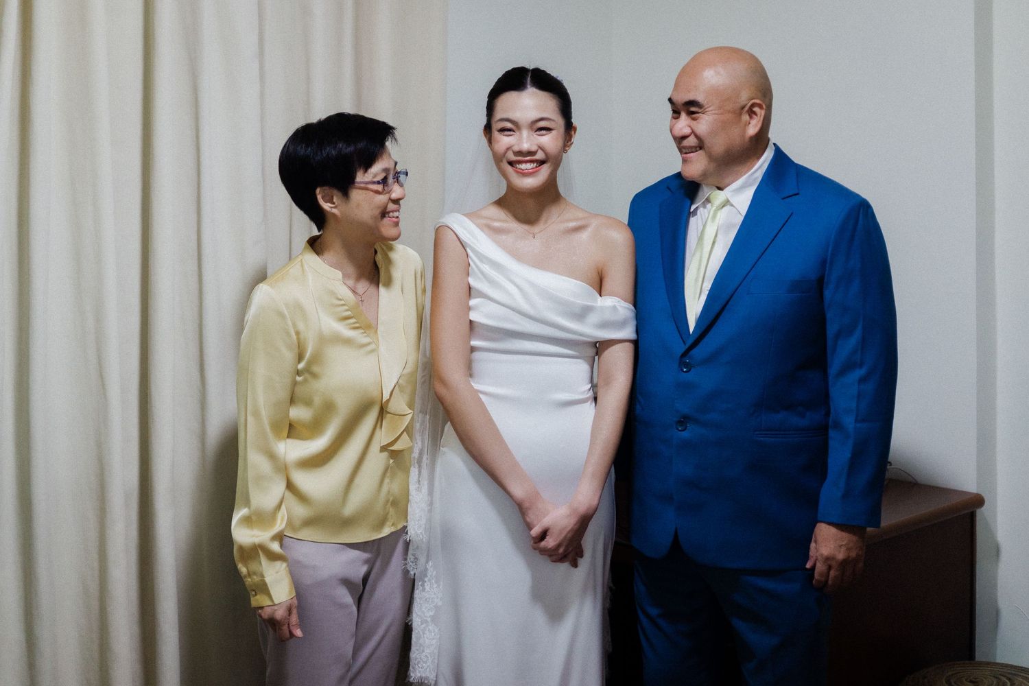 A bride in a white gown poses with two smiling guests, a woman in yellow and a man in a blue suit.