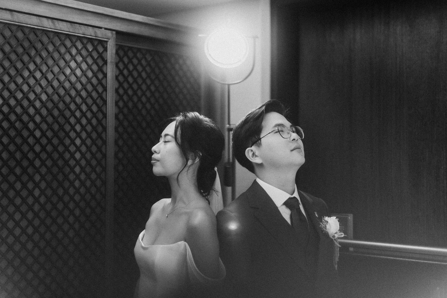 A black and white photo of a couple sitting back to back in formal attire at what appears to be a wedding venue.