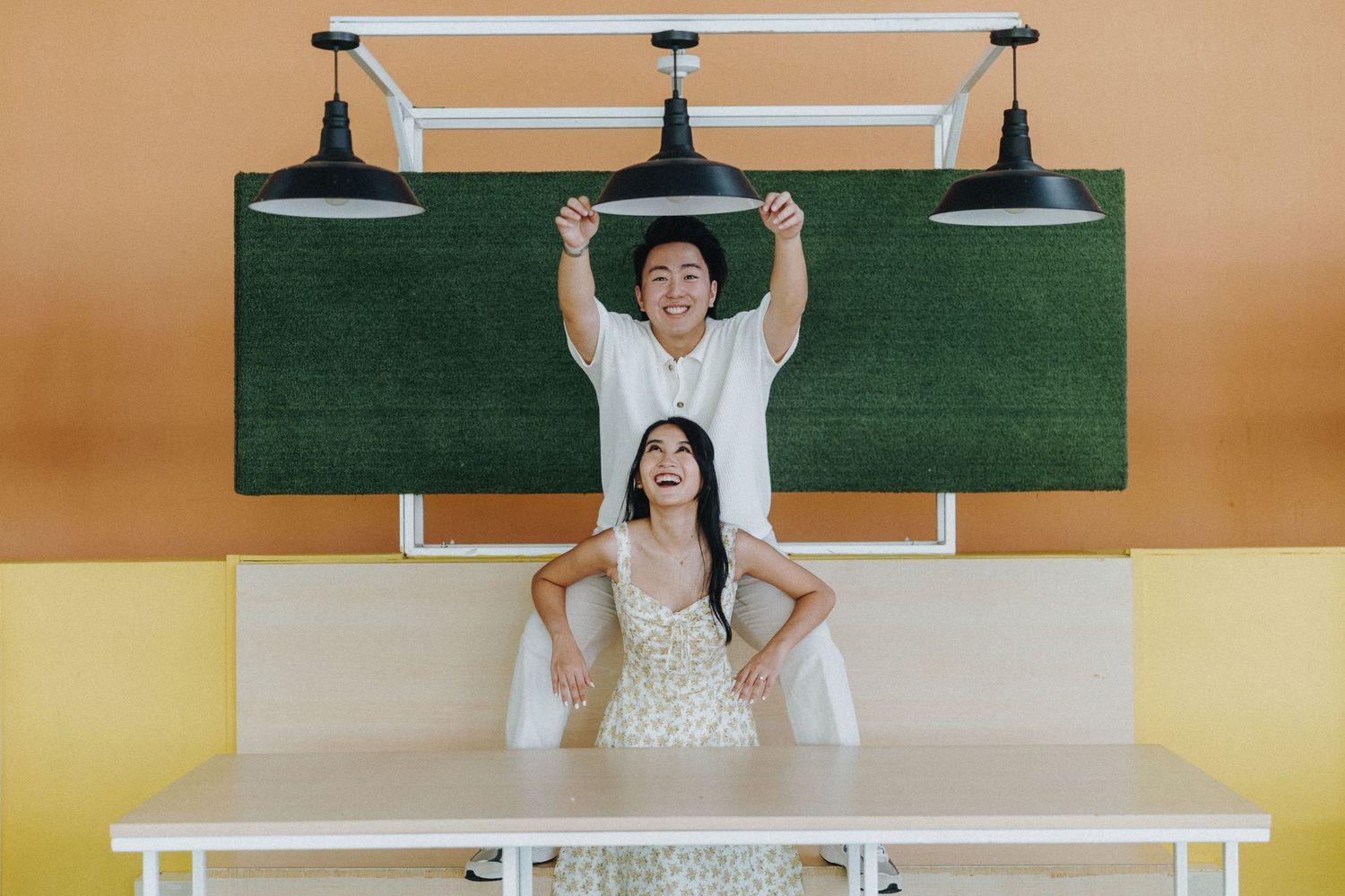 Playful couple pose against green chalkboard wall with pendant lights in minimalist setting.