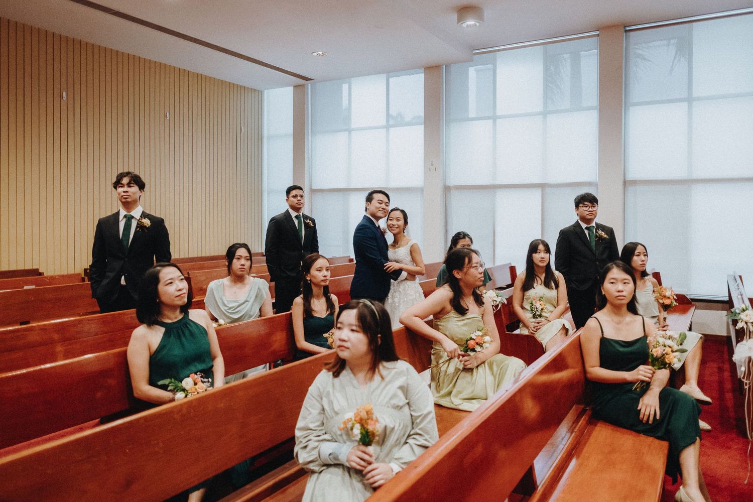 Wedding guests observe the ceremony from wooden pews in a bright, modern chapel with wood-paneled walls.