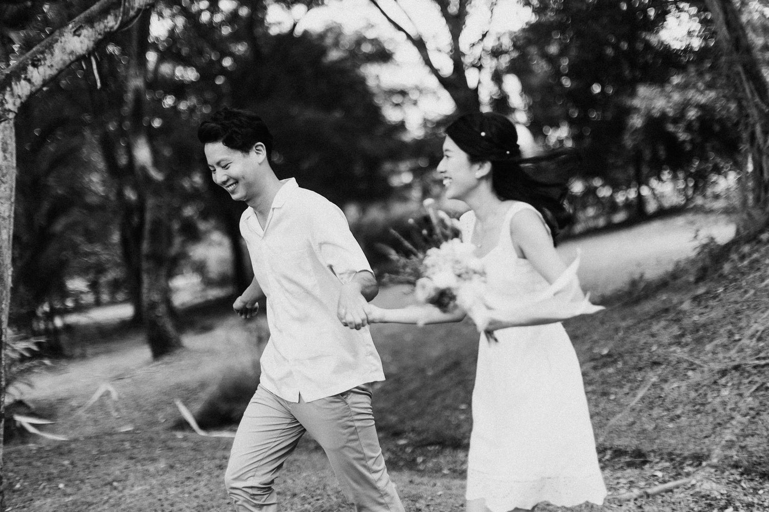 A couple in white clothing runs and laughs together on a hillside path in black and white.