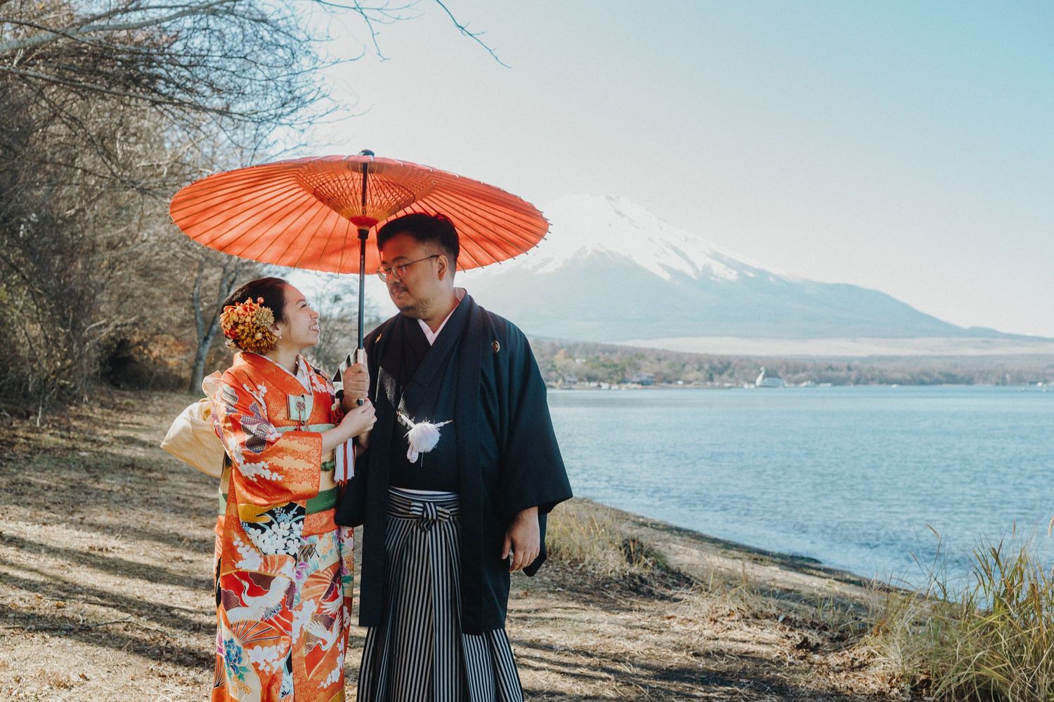 Traditional Japanese couple in kimono and hakama holding red umbrella with Mount Fuji view.