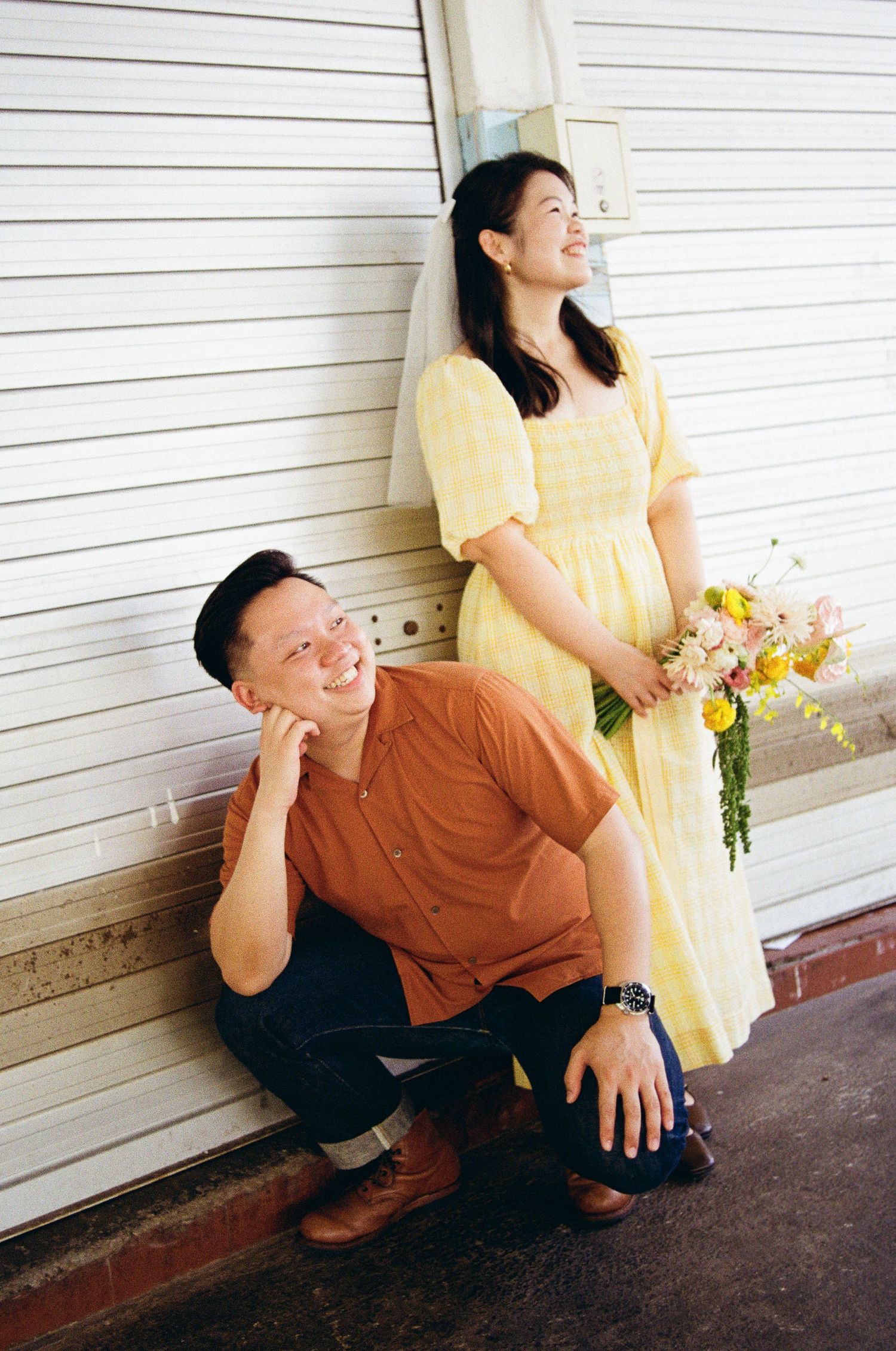 A couple in casual attire poses together against a white garage door, with one wearing a yellow dress.