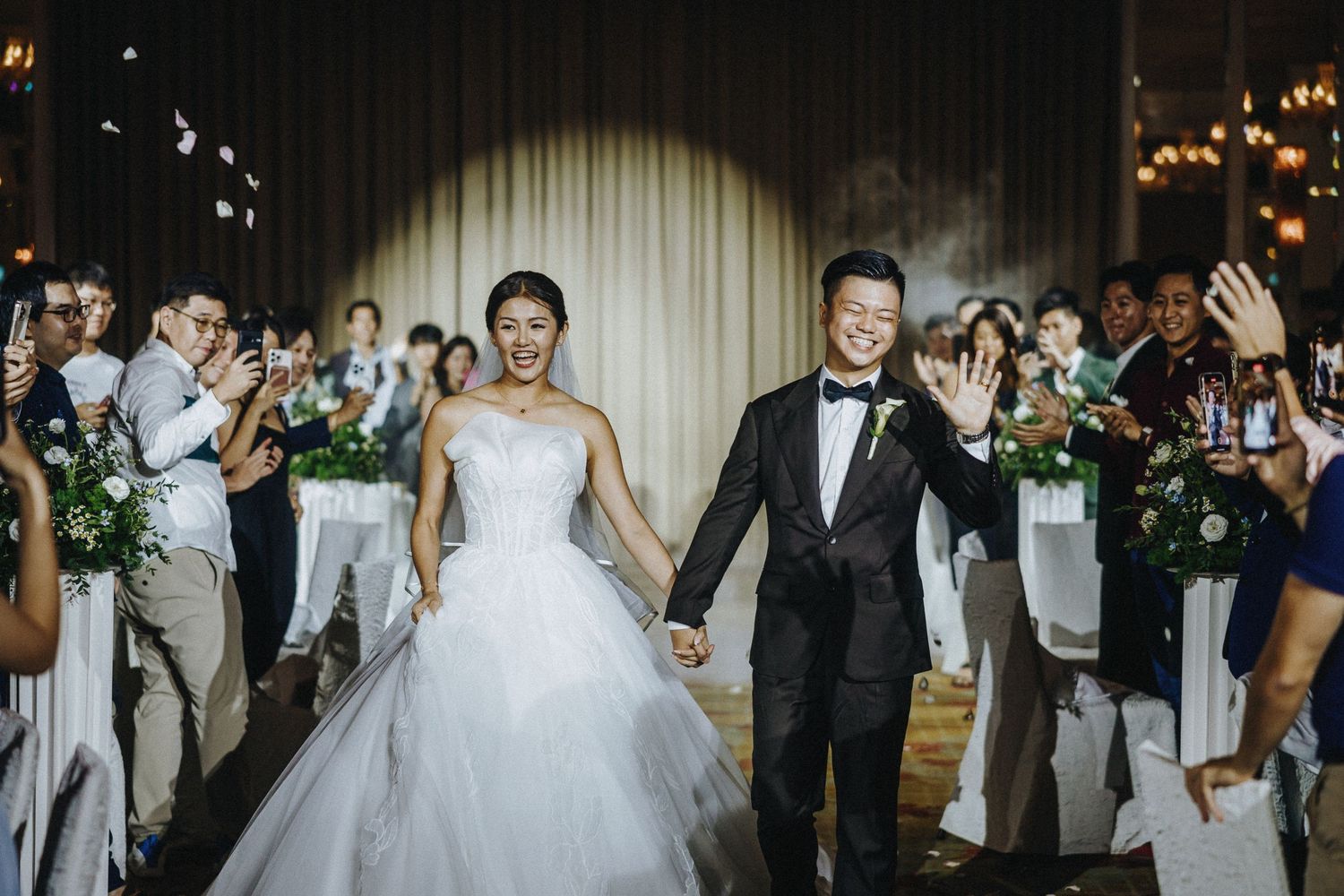 Newlyweds make their grand exit while wedding guests cheer and wave with sparklers in a dimly lit venue.
