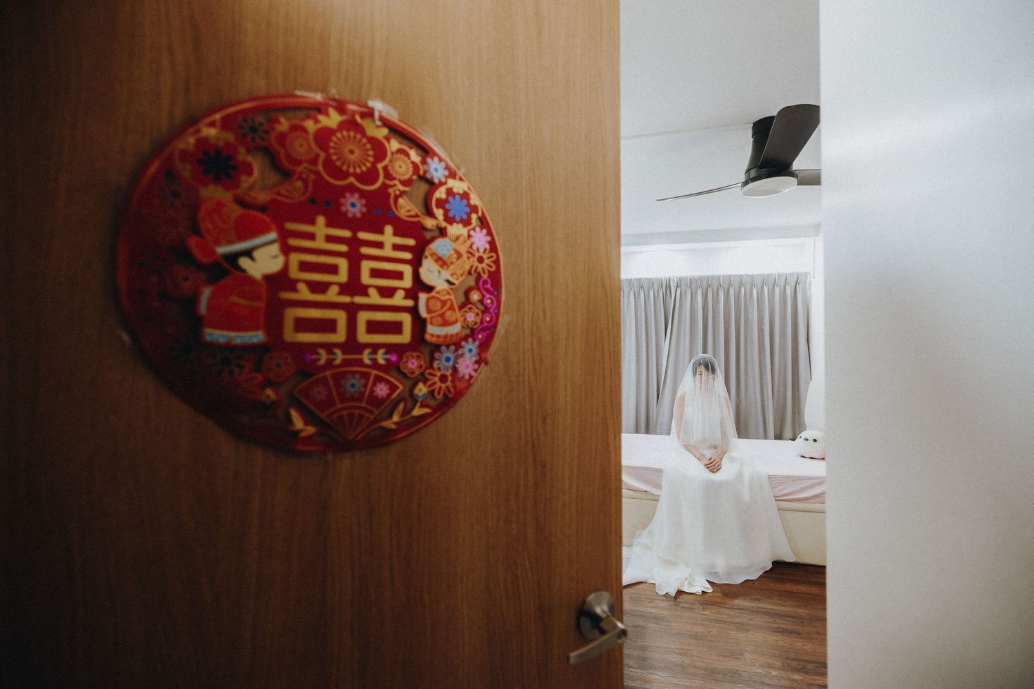 A traditional Chinese double happiness symbol decoration hangs on a door with a bridal preparation scene visible through doorway.