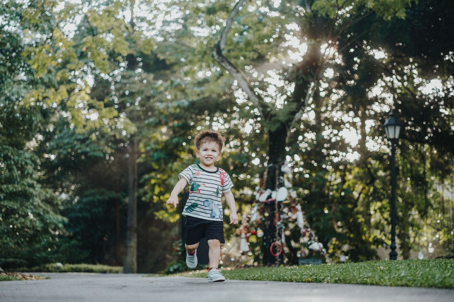Child in a striped shirt joyfully runs through a park on a sunny day with trees in the background.