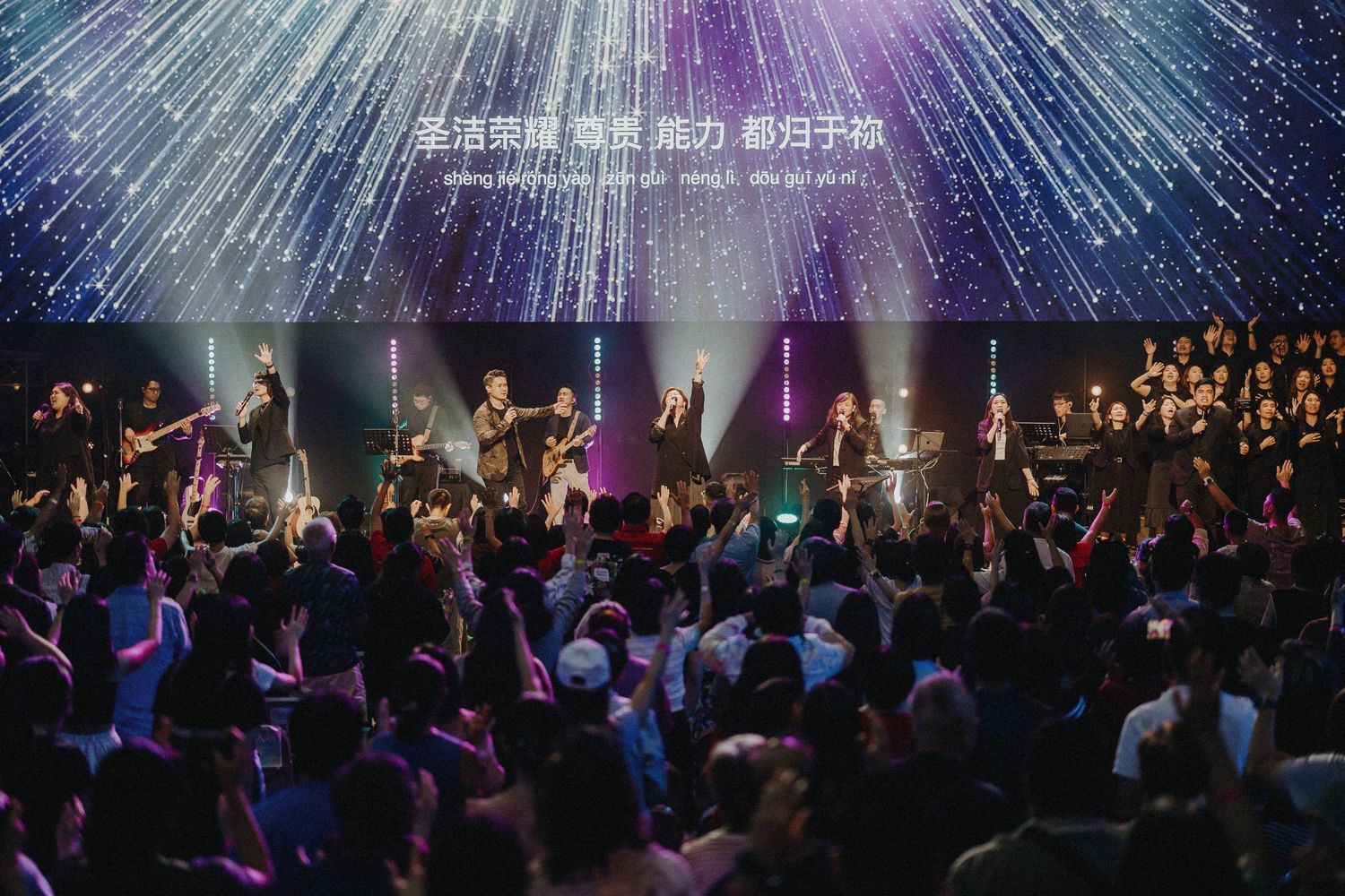 A large crowd worships with raised hands under dramatic purple and white lighting effects at a concert venue.