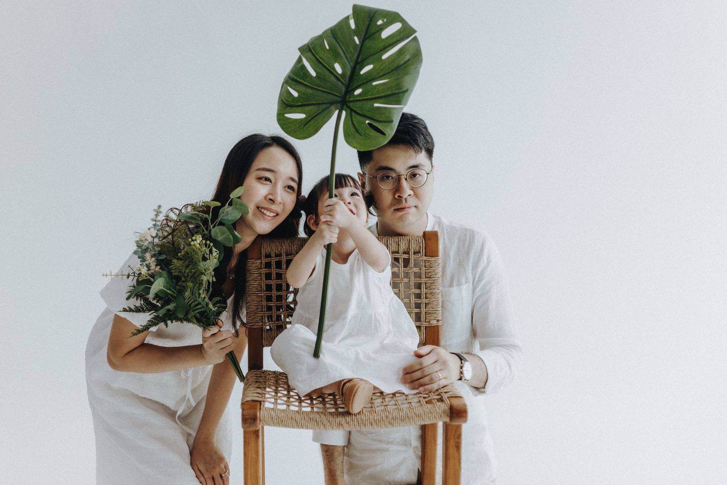 A minimalist family photoshoot in white against a light background with green monstera leaves.