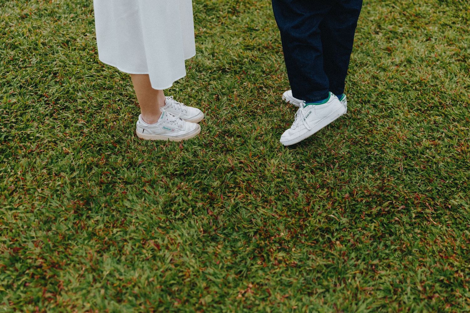 A row of people's legs and feet standing on green grass wearing white and blue sneakers and sandals.