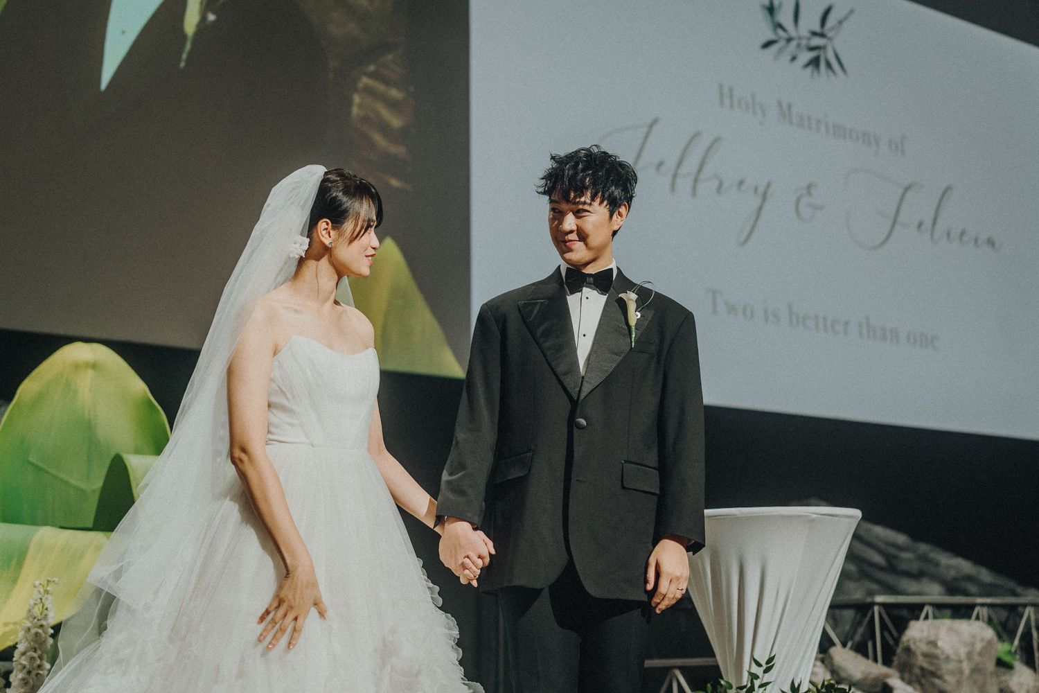 Wedding couple holding hands during ceremony with projection screen in background.