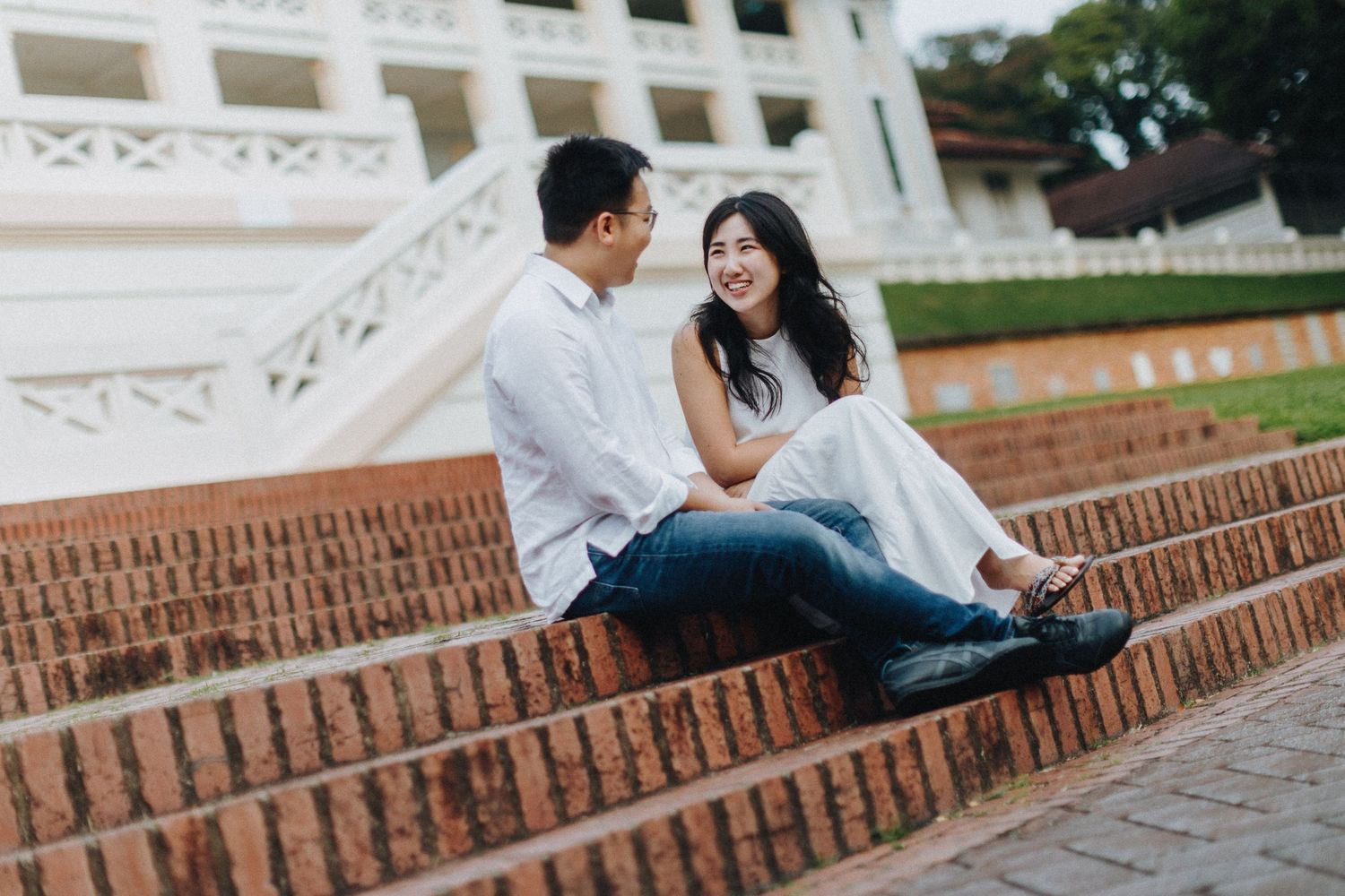 A couple in casual attire shares a romantic moment while sitting on brick steps at a historic building.