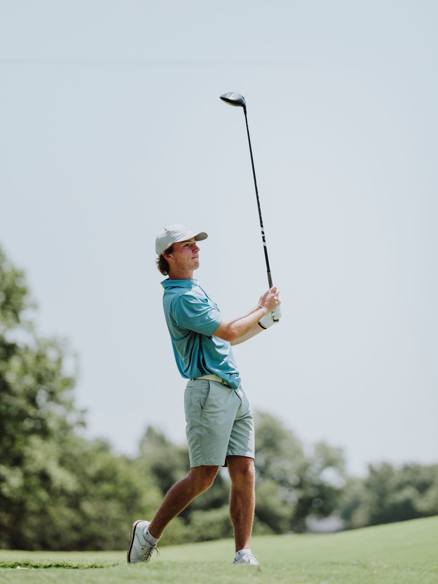 Golfer in teal polo shirt and light shorts follows through on swing during sunny day on golf course – Cedar Ridge golf photo by byDaviel.com