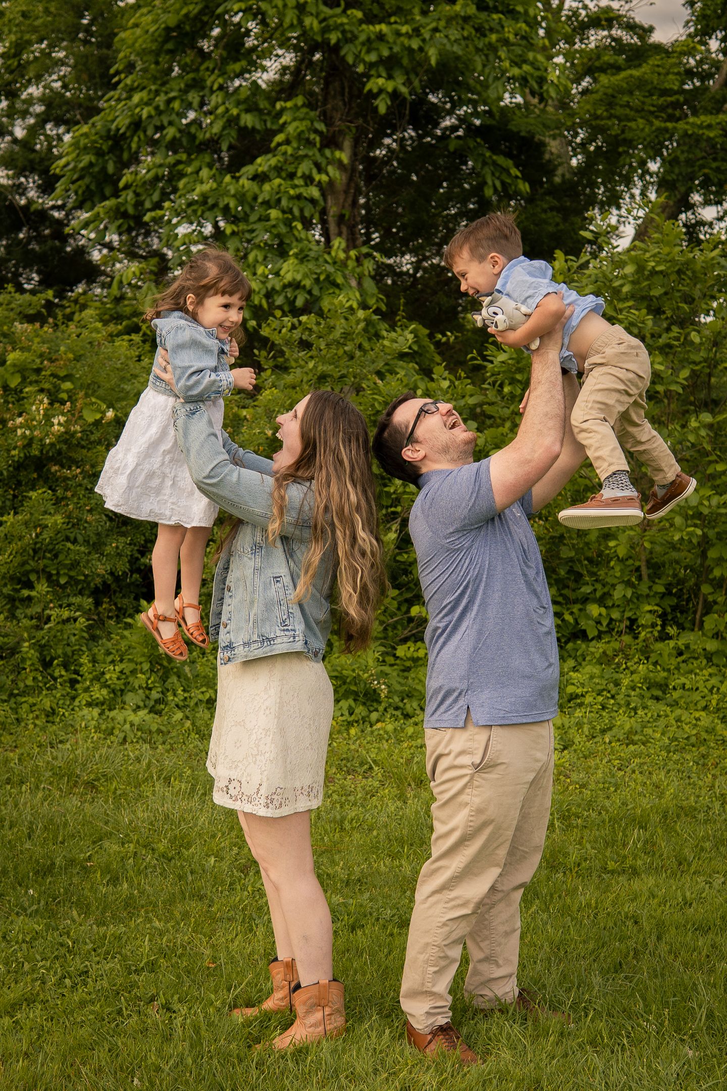 A family lifts two small children in the air while playing together in a lush green park setting.
