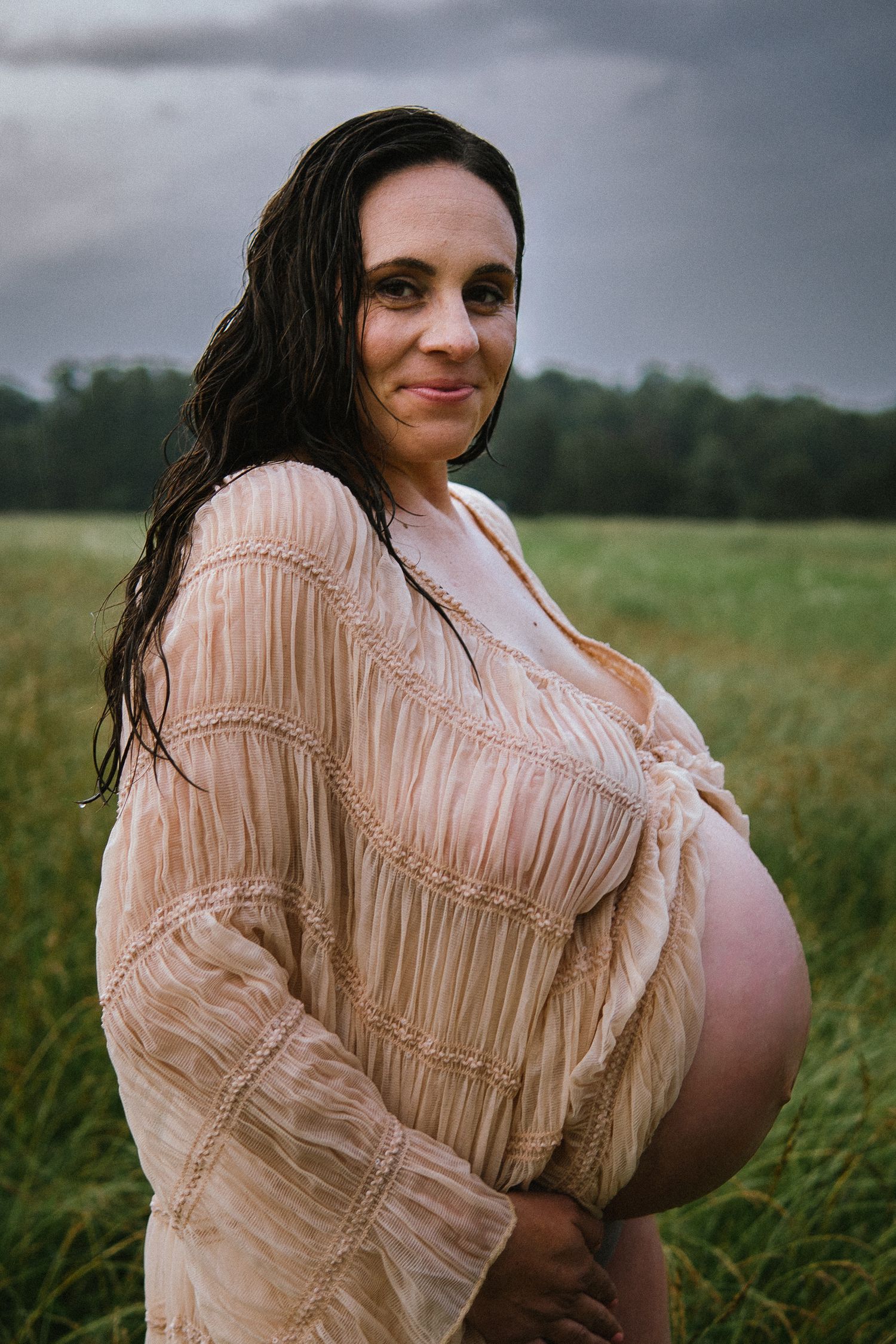 Maternity portrait of woman in vintage-style peach dress standing in natural field setting.