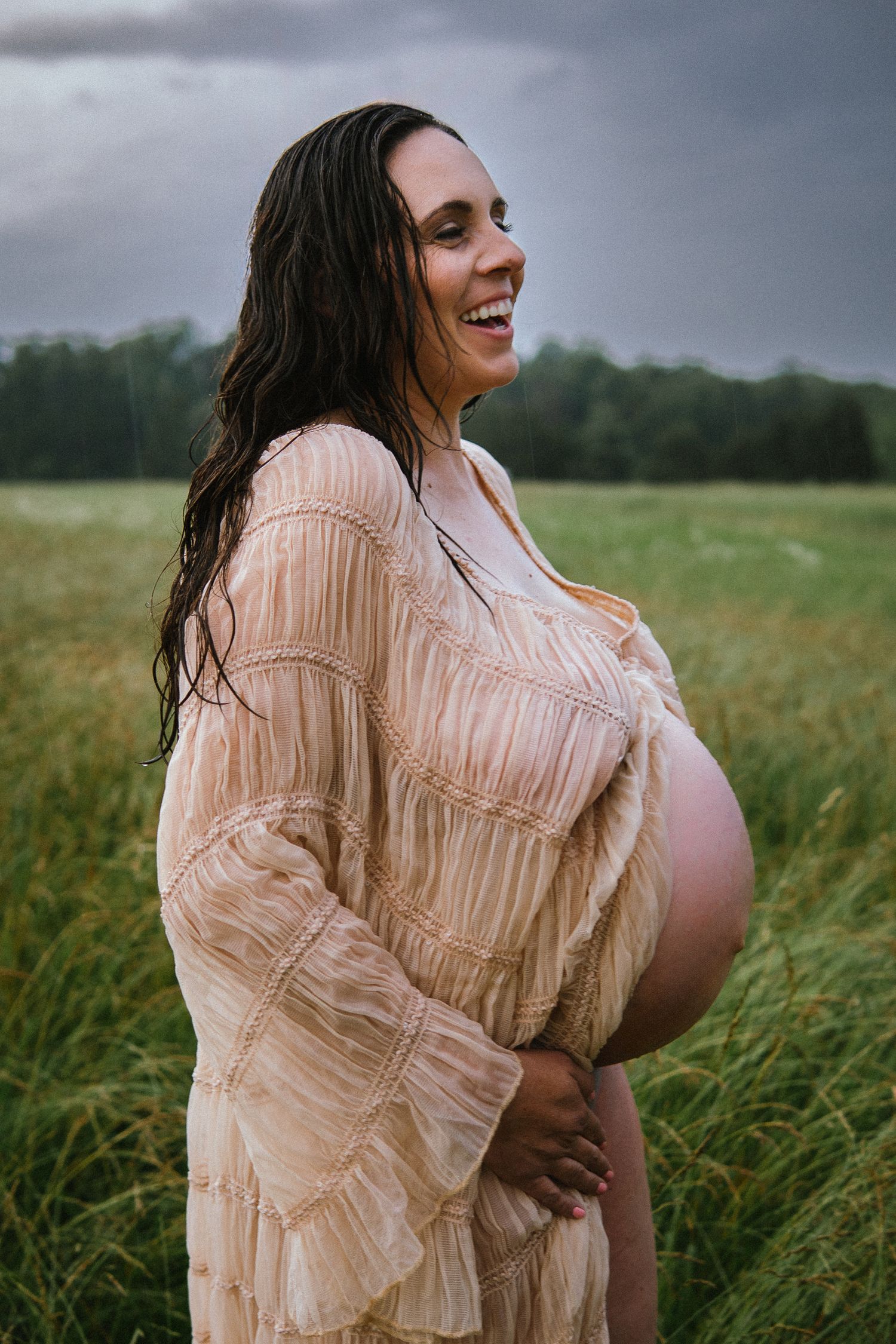 A joyful maternity portrait shows radiant laughter while wearing a romantic beige dress outdoors.