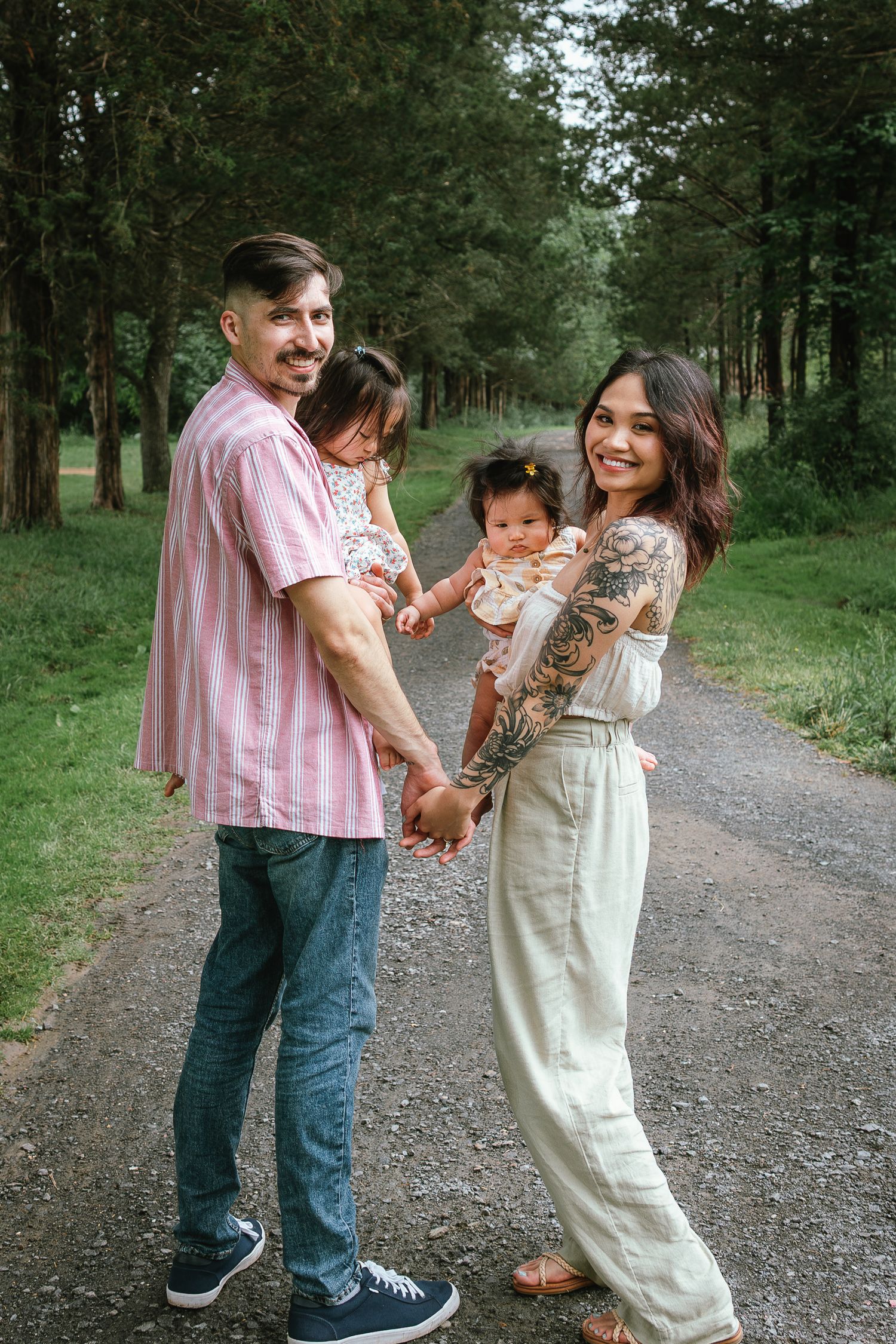 Family walks together on a tree-lined gravel path during a casual outdoor photo session.