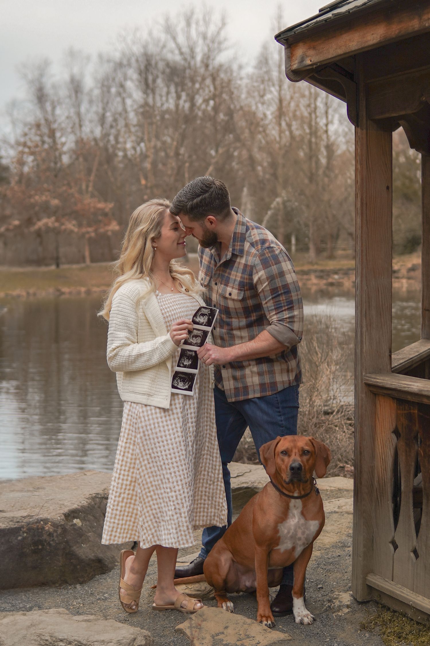 A couple shares an intimate moment by a lake with their brown dog while holding ultrasound photos in autumn.
